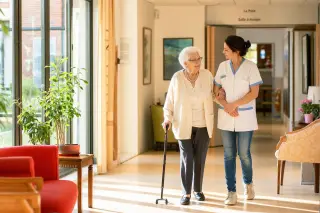 Attentive caregiver helps elderly resident walk in a bright, welcoming nursing home corridor with natural light and comfortable decor, both looking serene and dignified.