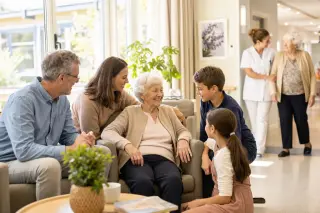 elderly resident with visiting family in a bright EHPAD living room, staff assisting another resident in the background, warm and dignified atmosphere